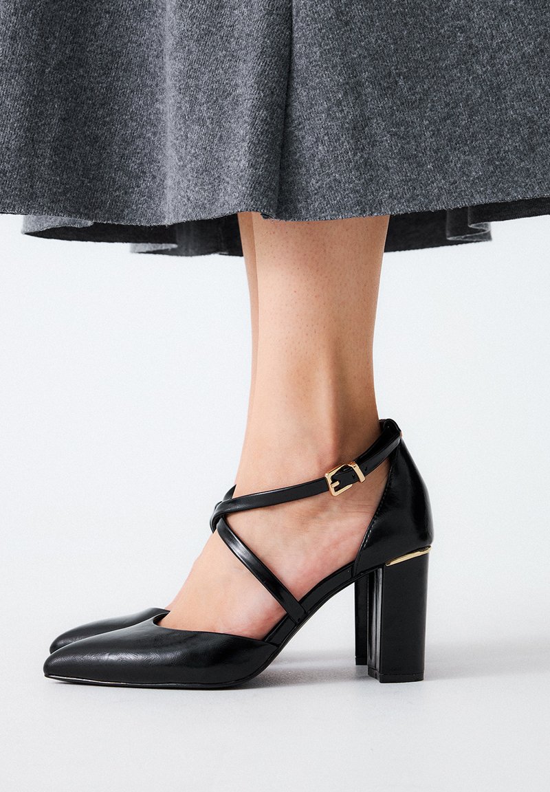 Zapatos de tacón alto de cuero negro con punta afilada, que cuentan con una correa entrelazada y un acento de hebilla dorada en la correa del tobillo.