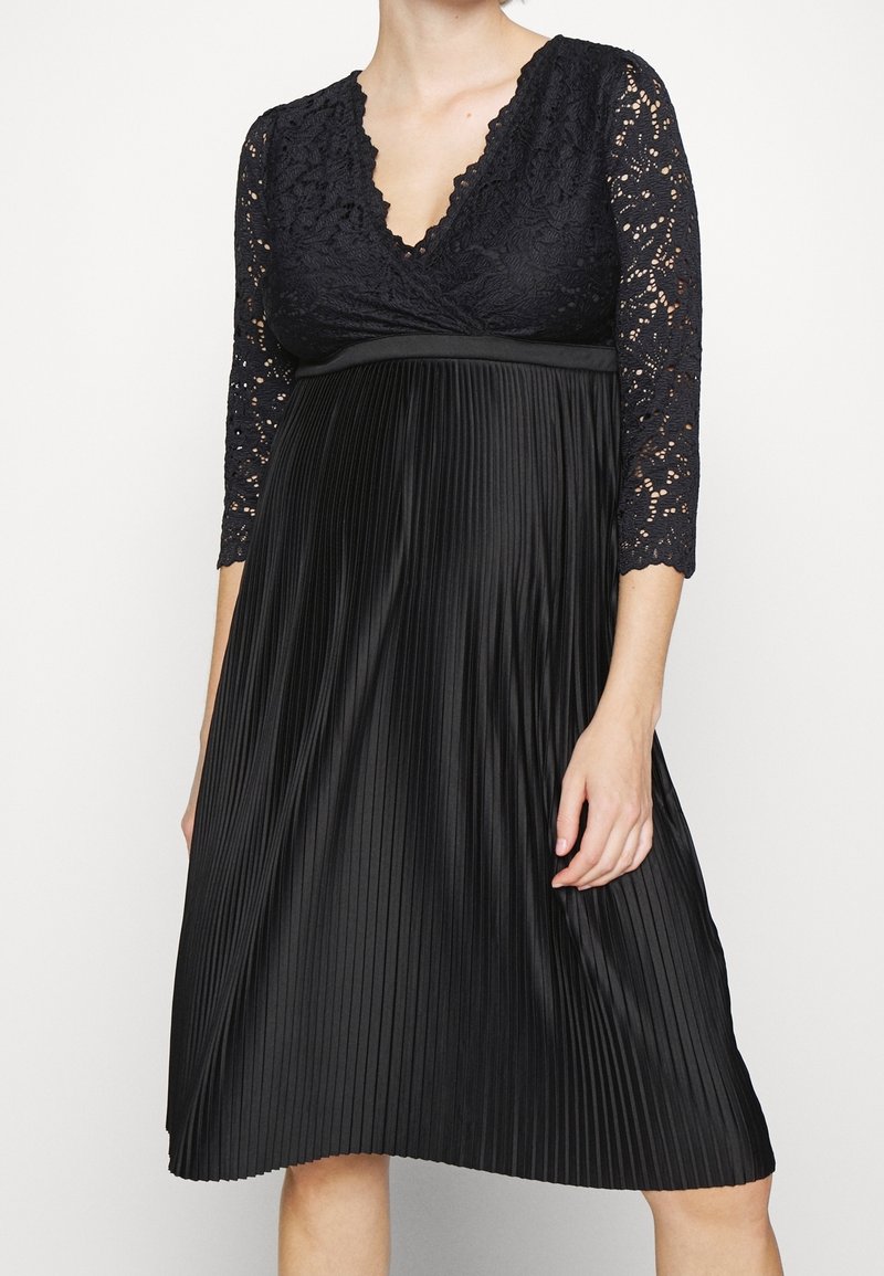 Robe noire avec un corsage en dentelle à encolure en V et des manches trois-quarts. La jupe plissée arrive au genou et est dotée d'une ceinture lisse.