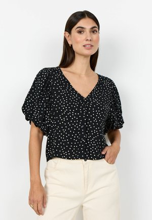 Woman wearing a black V-neck blouse with white polka dots and puffed short sleeves, paired with high-waisted white pants.