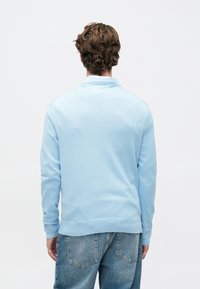 Barna hullámos hajú férfi világoskék pulóverben és kék farmernadrágban, hátulról, egy sima fehér háttér előtt.