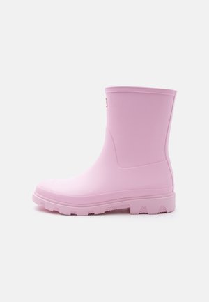 Solidní světle růžová gumová dešťová bota do poloviny lýtek s vroubkovanou podrážkou a hladkým povrchem, zobrazená z profilu na jednobílém pozadí.