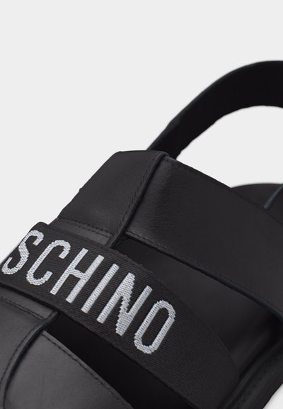 Μαύρο σανδάλι με χοντρές λουρίδες και φαρδιά ελαστική ταινία μπροστά με το κείμενο "SCHINO" σε λευκό.