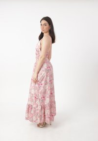Robe maxi en tissu rose avec motif floral, taille élastique, bretelles fines et ourlet volanté. Portée avec des sandales à talons, sur fond uni.
