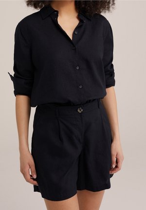 Kvinde iført sort langærmet skjorte med knapper og optrukne ærmer samt sorte shorts med høj talje og knaplukning.