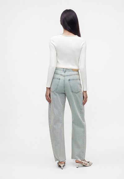 Femme aux longs cheveux noirs portant un haut blanc à manches longues, un jean large bleu clair et des chaussures blanches à talons bas, vue de dos sur fond blanc.