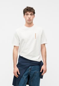 Νεαρός άνδρας με κατσαρά μαλλιά, φοράει λευκό t-shirt Timberland και μπλε τζιν, με μπλε σκούρο φούτερ δεμένο γύρω από τη μέση, στέκεται μπροστά σε λευκό φόντο.