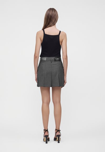 Femme portant un haut noir sans manches, une mini-jupe plissée grise avec patch d’étiquette et des sandales noires à talons, debout de dos.