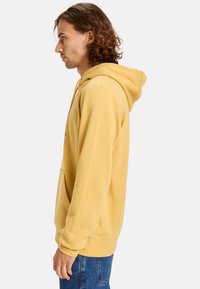 Sudadera amarilla con capucha, hecha de tejido suave, con un bolsillo tipo canguro y puños acanalados, combinada con jeans de mezclilla azules.