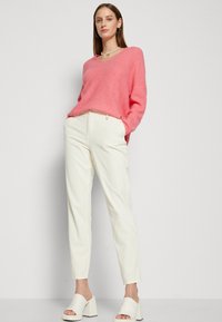 Ροζ πλεχτό πουλόβερ με V λαιμόκοψη, συνδυασμένο με κρέμ παντελόνι κομμένο κατά παραγγελία. Λευκά γόβες με τετράγωνη φτέρνα ολοκληρώνουν το σύνολο.