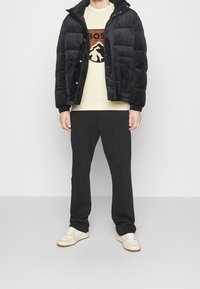 Black puffer jacket with a high collar, cream sweatshirt featuring a logo, black sweatpants, and beige sneakers with white accents.
