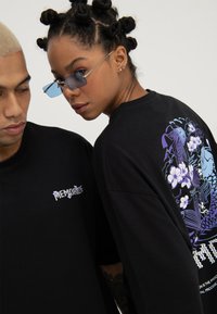 Zwei Personen tragen schwarze Shirts, eines mit dem Schriftzug "MEMORIZE", das andere mit einem violett-blauen Koi-Fisch- und Blumendesign auf der Rückseite.