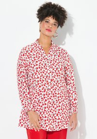 Blusa rossa e bianca con motivi a cuore, maniche lunghe, colletto, chiusura a bottoni sul davanti e orlo leggermente svasato. I bottoni sono dello stesso colore della blusa.
