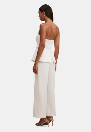 Kvinne iført en stroppeløs hvit peplumtopp og vide hvite bukser, stående sidelengs, med gullfargede høye hæler og øredobber.