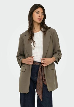 Ung kvinde med langt mørkt hår iført en åben taupe blazer, hvid tekstureret top, mørke jeans og et brunt mønstret tørklæde som bælte.