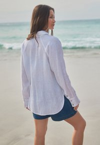 Blouse en lin blanc à manches longues, poignets boutonnés et coupe décontractée. Associée à un short en jean, sur fond de plage.