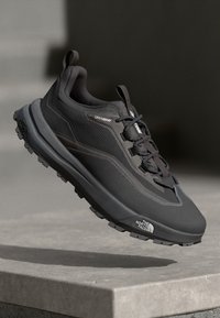 Zapato deportivo negro con una superficie texturizada, que presenta materiales de malla y sintéticos, una suela acolchada y un sistema de cordones. El logo de la marca es visible.