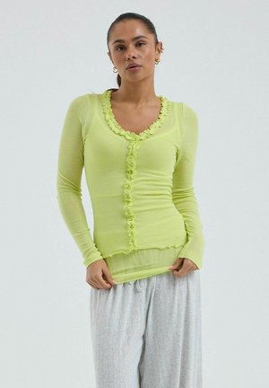 Žena so stiahnutými vlasmi nosí priehľadný limetkovo zelený dlhý rukáv s volánikovým lemom a svetlo sivé voľné nohavice na jednofarebnom pozadí.