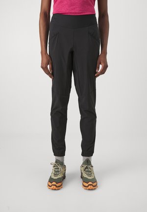 Pantaloni sportivi neri con una superficie liscia, gamba affusolata e polsini elasticizzati. Due tasche laterali angolate; indossati con scarpe robuste e multicolori.