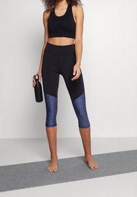 Soutien-gorge de sport noir associé à des leggings capri noirs avec un motif moucheté bleu et une texture lisse. Le modèle tient une bouteille d'eau en argent.