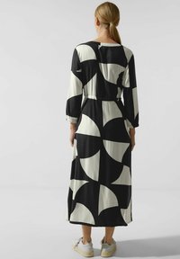 Kobieta widziana z tyłu, ubrana w długą sukienkę w czarno-białe geometryczne wzory z rękawami 3/4 i białymi sneakersami, stojąca na szarym podłożu.