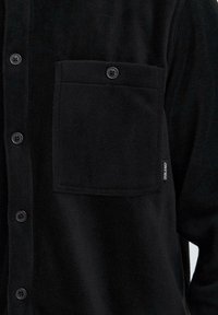 Black fleece shirt with button closure, large chest pocket, and subtle branding tag; soft texture and smooth finish.