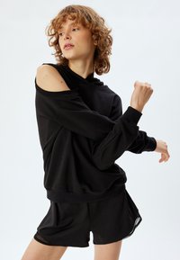 Sudadera negra con diseño off-the-shoulder y mangas largas, combinada con pantalones cortos negros con un dobladillo translúcido.
