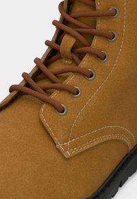 Beżowe zamszowe botki z okrągłym noskiem, brązowymi sznurowadłami, metalowymi oczkami, kontrastowym przeszyciem i czarną gumową podeszwą. Teksturowana powierzchnia i gładkie wykończenie.