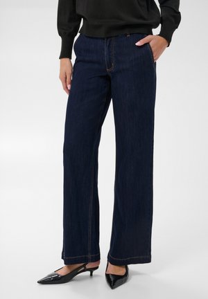 Femme portant un jean bleu foncé à jambes larges avec un pull noir, une main dans la poche et des chaussures noires pointues à talons bas.