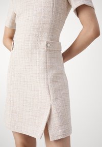 Femme portant une robe beige texturée à manches courtes, avec une fente sur le côté et des détails de ceinture boutonnée, se tenant les mains derrière le dos.
