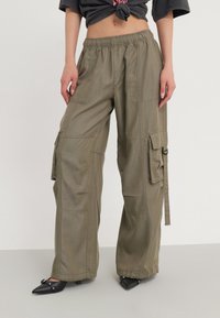 Olivgrüne Cargo-Hosen mit lockerer Passform, elastischem Bund, mehreren Taschen und leichtem Stoff, kombiniert mit schwarzen, spitz zulaufenden Pumps.