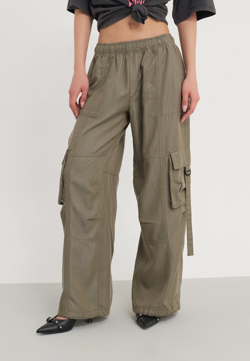 Olivgrüne Cargo-Hosen mit lockerer Passform, elastischem Bund, mehreren Taschen und leichtem Stoff, kombiniert mit schwarzen, spitz zulaufenden Pumps.