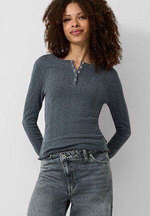 Frau mit lockigem Haar, die ein langärmeliges dunkelgraues Henley-Oberteil und hoch taillierte, ausgewaschene graue Jeans trägt, steht vor einem schlichten Hintergrund.
