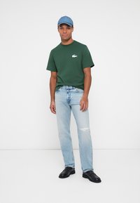 Tricou verde din bumbac cu un mic logo alb, blugi din denim bleu deschis cu detalii distorsionate și pantofi din piele negri. Capp bleu deschis.