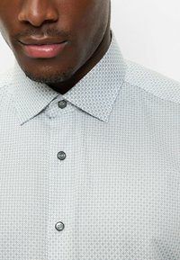 Hellblaues Hemd mit Knopfleiste und geometrischem Muster. Es hat einen spitzen Kragen und zwei dunkle Knöpfe, gefertigt aus einem glatten Stoff.