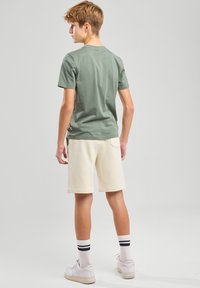 Camiseta de algodón verde, de manga corta, combinada con pantalones cortos color crema. Zapatillas blancas y calcetines a rayas completan el atuendo. Diseño simple y casual.