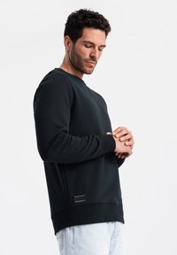 Sudadera negra de mezcla de algodón; presenta puños y cuello de canalé, un corte recto y una etiqueta lateral. El modelo lleva puesta unos vaqueros de color azul claro.