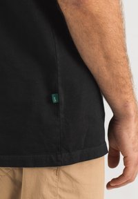 Schwarzes Baumwoll-T-Shirt mit seitlichem Schlitz, ausgestattet mit einem grünen Stoffetikett. Glatte Textur, gerader Schnitt und dezente Nähte am Saum.