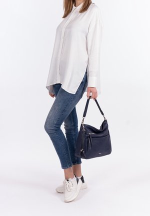 Granatowa skórzana torebka z srebnymi akcentami, z zamkiem błyskawicznym oraz zamykanymi kieszeniami. Noszona z białą koszulą na guziki i niebieskimi dżinsami.