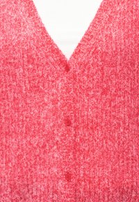 Κόκκινη πλεκτή ζακέτα με υφή, V-λαιμόκοψη και τρία ταιριαστά κουμπιά μπροστά, φορεμένη πάνω από λευκό τοπ.