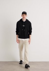 Sudadera negra con un logo, superpuesta sobre una camiseta blanca, combinada con pantalones beige y zapatos negros. Lleva una gorra negra. Fondo neutro.