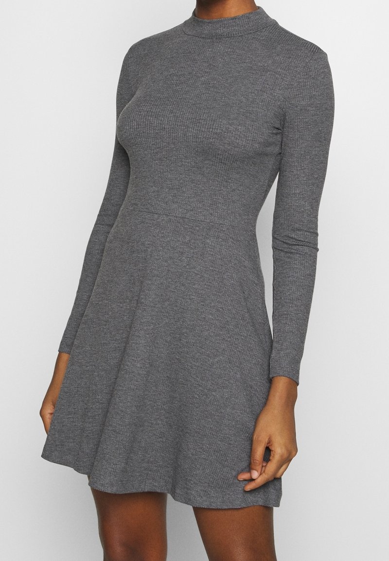 Femme portant une robe grise moulante à manches longues, en maille côtelée, avec un col montant et une jupe évasée sur un fond clair uni.