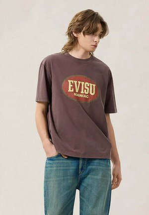 Giovane con capelli di media lunghezza che indossa una t-shirt marrone EVISU MANIAC e jeans blu, in piedi con una mano in tasca.