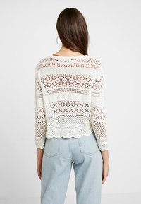 Maglione bianco all'uncinetto con orlo a scallop, caratterizzato da motivi a rete e maniche lunghe, abbinato a jeans azzurri a vita alta.