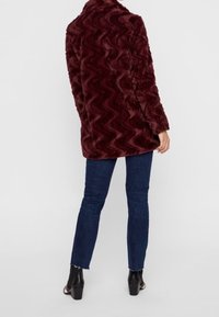 Burgundowa kurtka z sztucznego futra o falistej fakturze, z kołnierzem i lekko oversize'owym krojem, w połączeniu z ciemnymi obcisłymi dżinsami i botkami za kostkę.