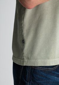Licht olijfgroen katoenen T-shirt met zijnaaddetails en een klein zwart label aan de linkerkant. Draagt met donkere spijkerbroek.
