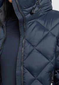 Chaqueta de plumas acolchada de color azul marino con un alto cuello, cierre frontal con cremallera y un brillo sutil. Los detalles incluyen un cordón y herrajes de cremallera en negro.
