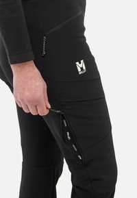 Pantalons softshell noirs avec poches zippées, offrant une coupe près du corps et un tissu texturé, ornés d'un logo Millet blanc sur la cuisse.