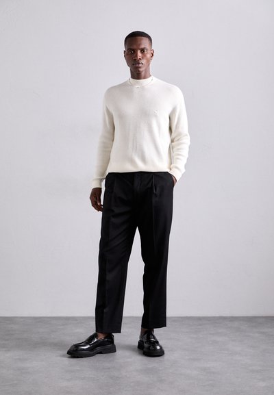 Pulover crem cu nervuri, pantaloni negri adaptați și loaferi negri lucioși. Design simplu cu detalii subtile, pe un fundal neutru.