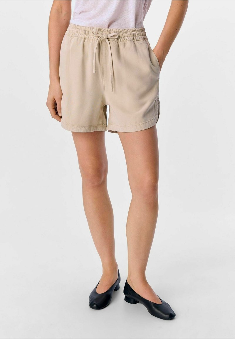 Femme portant un short beige à cordon de serrage avec poches latérales et des chaussures noires à talons bas sans lacets, debout, une main dans la poche.
