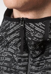 Close-up of a person’s neck wearing a black and white patterned zip-up garment with a large black zipper pull.